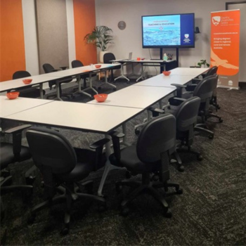 Training room with U-shaped tables, black chairs, a large screen displaying a presentation, and an orange banner in the corner.